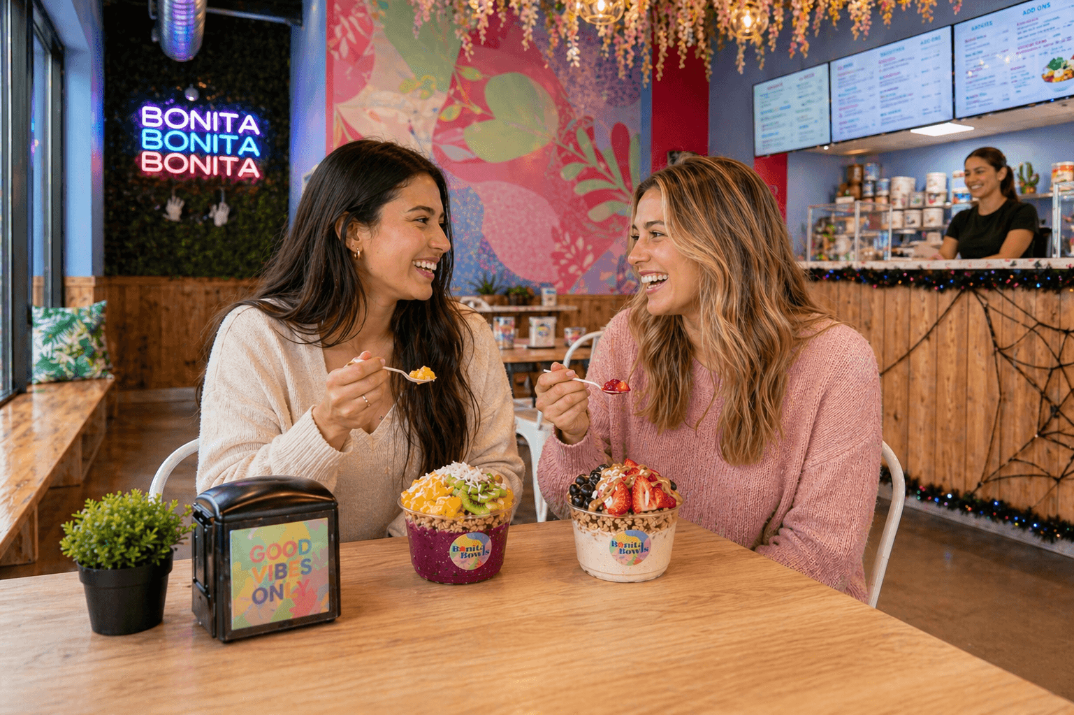 Two friends sharing açaí bowls inside a Bonita Bowls shop, BONITA neon sign and flower wall in the background